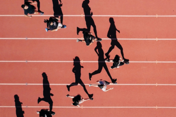 Aerial view of a group of people running on a red track with white lane markings. Their long shadows stretch across the lanes, creating a dynamic and engaging visual. The image captures movement and competition, symbolizing progress and the race to stay ahead.