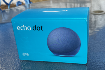 A blue box featuring Amazon's Echo Dot smart speaker is placed on a reflective outdoor table. The packaging prominently displays an image of the spherical Echo Dot in blue, along with the words "echo dot" and the Alexa logo. The background includes a blurred outdoor setting with a blue metal chair and part of a yard visible.